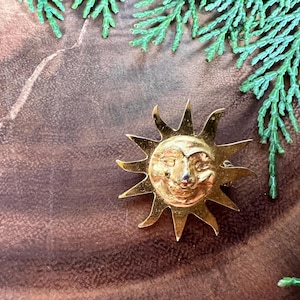 May include: A gold-toned sun pin with a face and a textured surface. The pin is on a brown wooden surface.