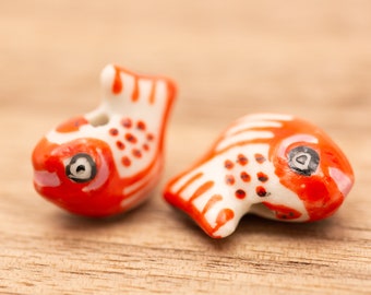 May include: Two small ceramic fish charms, one red and white with black eyes and the other white and red with black eyes. The fish are on a wooden surface.