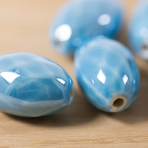 May include: Five smooth, oval-shaped ceramic beads in a light blue color with a marbled pattern. The beads have a glossy finish and a hole through the center.