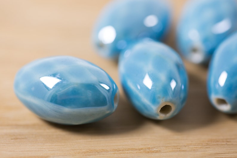 May include: Five smooth, oval-shaped ceramic beads in a light blue color with a marbled pattern. The beads have a glossy finish and a hole through the center.