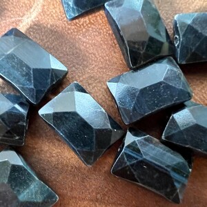 May include: A collection of dark blue faceted gemstone beads. The beads are rectangular in shape and have a smooth, polished finish.