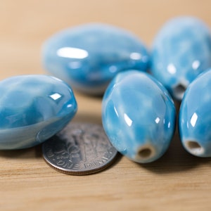 May include: A group of six light blue ceramic oval beads with a white swirl pattern. The beads are smooth and glossy.