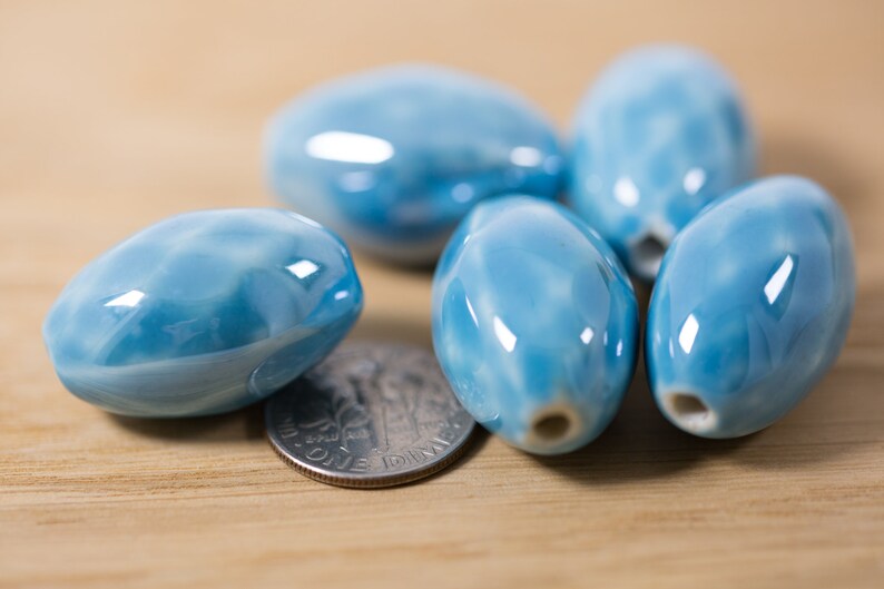 May include: A group of six light blue ceramic oval beads with a white swirl pattern. The beads are smooth and glossy.