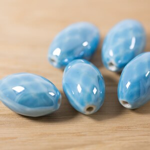 May include: Six light blue ceramic oval beads with a marbled effect. The beads are smooth and glossy.