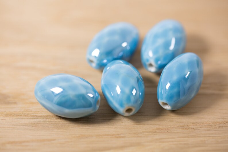 May include: Six light blue ceramic oval beads with a marbled effect. The beads are smooth and glossy.