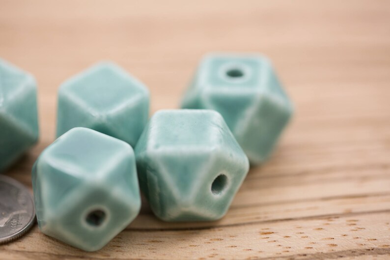 May include: A group of six teal blue ceramic beads with a geometric shape. The beads have a smooth, glazed finish and a hole in the center for stringing.