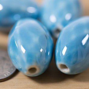 May include: A close-up of several light blue ceramic oval beads with a white marbled pattern. The beads are smooth and glossy.