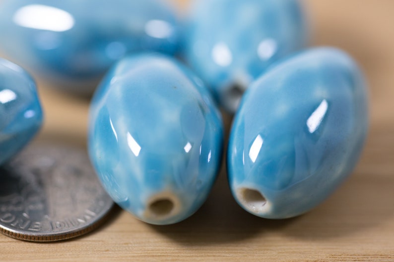 May include: A close-up of several light blue ceramic oval beads with a white marbled pattern. The beads are smooth and glossy.