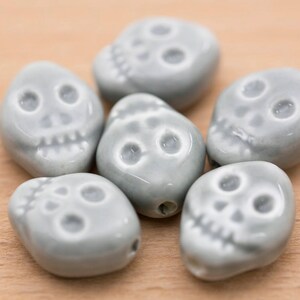 May include: Six grey ceramic skull beads with white detailing. The beads are smooth and have a slightly glossy finish.