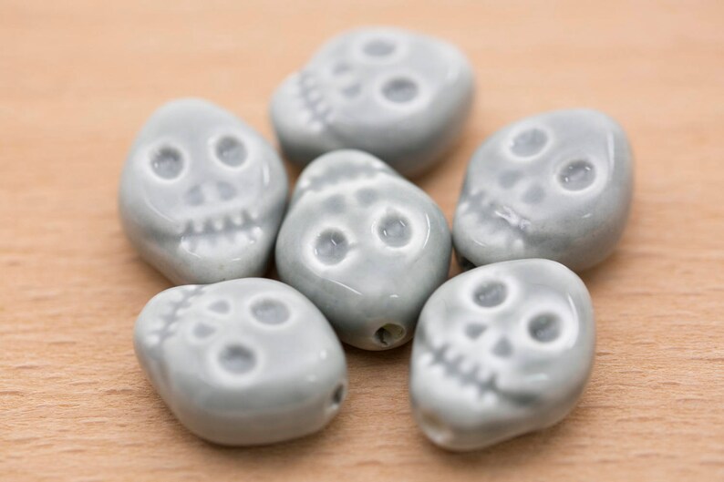 May include: Six grey ceramic skull beads with white detailing. The beads are smooth and have a slightly glossy finish.