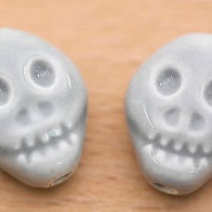 May include: Two ceramic skull beads in a light grey colour. The beads have a simple design with two eyes, a nose, and a mouth.