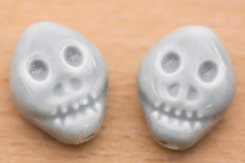 May include: Two ceramic skull beads in a light grey colour. The beads have a simple design with two eyes, a nose, and a mouth.