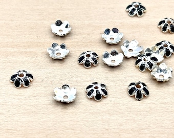 May include: A group of silver-toned flower-shaped bead caps with black enamel accents. The caps are scattered on a light brown wooden surface.