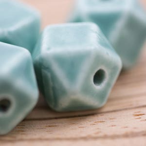 May include: Four teal blue ceramic beads with a geometric shape and a hole in the center. The beads are on a light brown wooden surface.