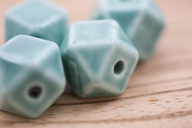 May include: Four teal blue ceramic beads with a geometric shape and a hole in the center. The beads are on a light brown wooden surface.