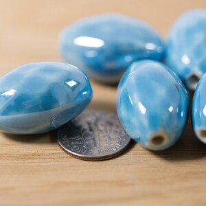 May include: A group of six oval-shaped ceramic beads in a light blue color with white swirls. The beads are smooth and glossy, and they are arranged in a cluster on a wooden surface.