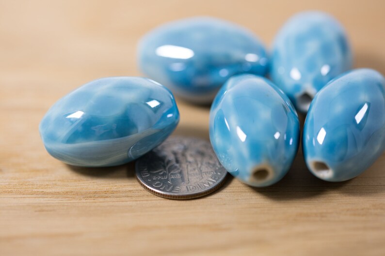 May include: A group of six oval-shaped ceramic beads in a light blue color with white swirls. The beads are smooth and glossy, and they are arranged in a cluster on a wooden surface.