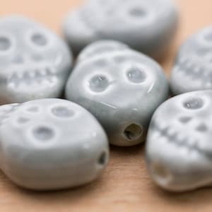May include: Six grey ceramic skull beads with white painted details. The beads are smooth and have a slightly glossy finish.