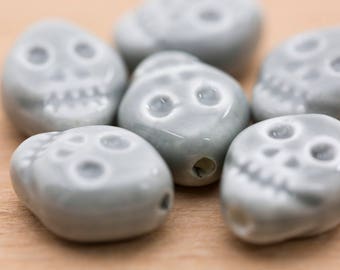 May include: Six grey ceramic skull beads with white painted details. The beads are smooth and have a slightly glossy finish.