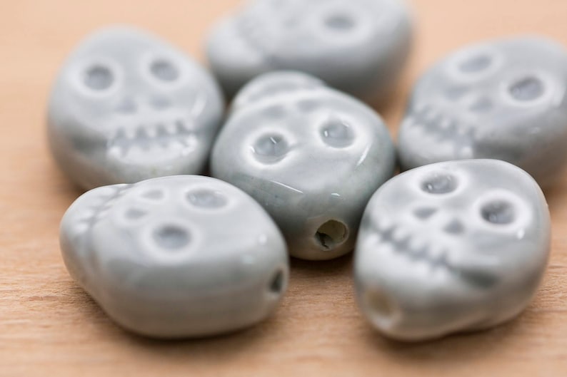May include: Six grey ceramic skull beads with white painted details. The beads are smooth and have a slightly glossy finish.