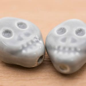 May include: Two grey ceramic skull beads with white details. The beads are smooth and have a glossy finish.