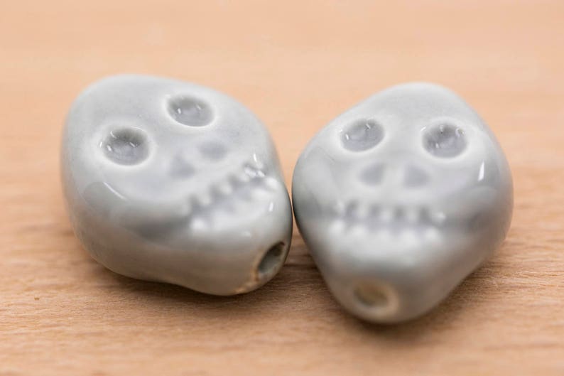 May include: Two grey ceramic skull beads with white details. The beads are smooth and have a glossy finish.