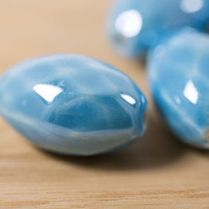 May include: A close-up of four light blue ceramic oval beads. The beads have a glossy finish and a subtle marbled pattern.