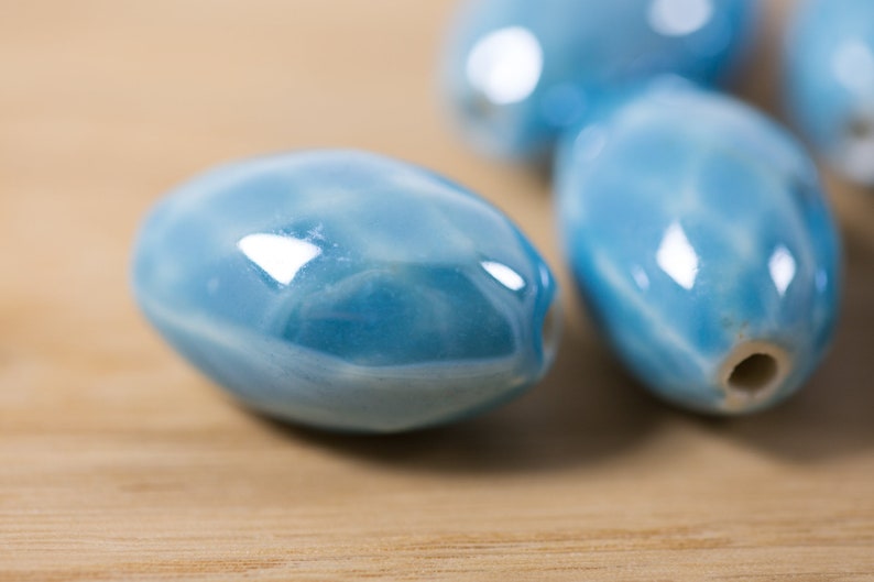 May include: A close-up of four light blue ceramic oval beads. The beads have a glossy finish and a subtle marbled pattern.