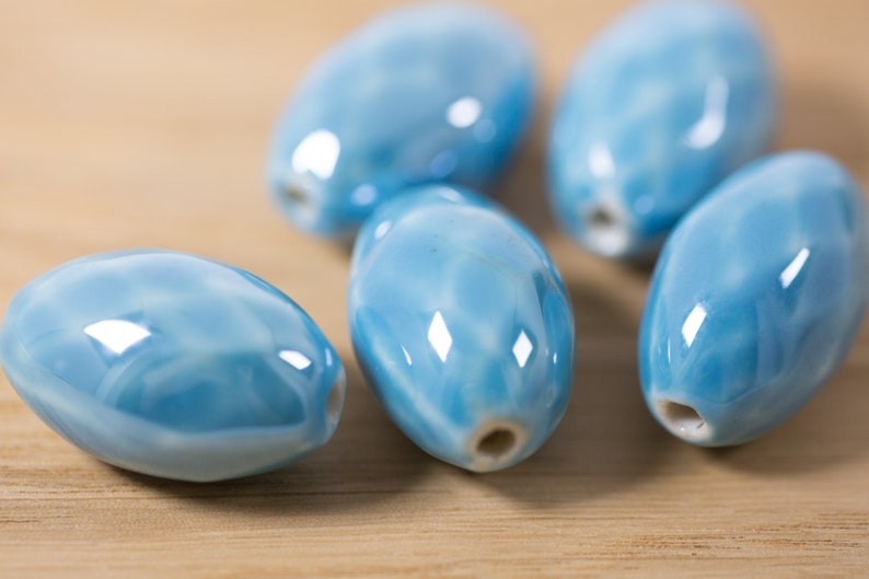 May include: Six light blue ceramic oval beads with a white marbled pattern. The beads have a hole through the center for stringing.