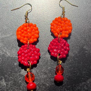 Puede incluir: Un par de pendientes colgantes con un diseño de tres niveles. El nivel superior es naranja, el nivel intermedio es rojo y el nivel inferior presenta cuentas rojas. Los pendientes tienen ganchos y herrajes de conexión dorados. El fondo es una superficie oscura y texturizada.
