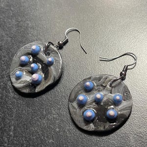 Puede incluir: Un par de pendientes hechos a mano. Cada pendiente es un disco circular con un patrón veteado en negro y gris. Pequeñas cuentas azules y blancas están dispersas por la superficie. Los pendientes tienen ganchos para las orejas de color plateado.