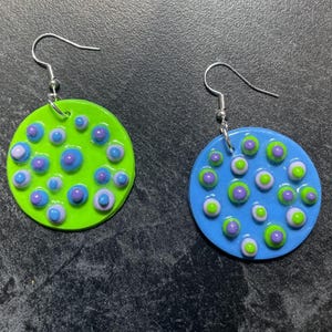 Puede incluir: Un par de pendientes hechos a mano. Uno es verde lima, el otro azul. Ambos son circulares con un patrón de puntos azules, morados y blancos en relieve. Los pendientes tienen ganchos plateados.