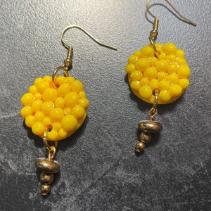 Puede incluir: Un par de pendientes hechos a mano con un grupo de pequeñas cuentas amarillas en forma circular. Los pendientes tienen ganchos dorados y una cuenta metálica texturizada colgante. El fondo es una superficie oscura y texturizada.