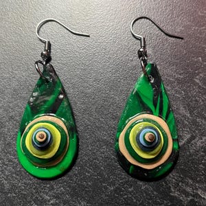 Puede incluir: Un par de pendientes hechos a mano con forma de lágrima. Los pendientes presentan un fondo veteado verde y negro con un diseño circular en capas en el centro. El diseño circular incluye tonos de verde, amarillo, azul y beige. Los ganchos de los pendientes son plateados.