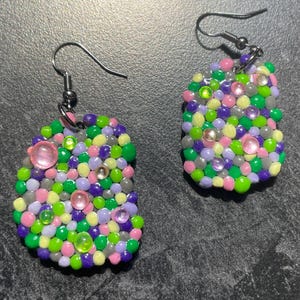 Puede incluir: Un par de pendientes hechos a mano. Cada pendiente es un grupo de pequeñas cuentas de colores en tonos verdes, morados, rosas y amarillos. Gemas claras y rosas se intercalan en el diseño. Los pendientes tienen ganchos plateados.