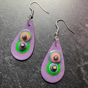Puede incluir: Un par de pendientes artesanales en forma de lágrima. Cada pendiente es de color lavanda con dos círculos coloridos apilados. Los círculos presentan detalles verdes, rosas y negros. Los pendientes tienen ganchos plateados.