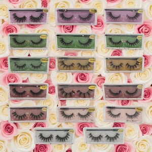 May include: A collection of false eyelashes in clear plastic cases, each labeled with a different name. The lashes vary in style and color, with some featuring glitter accents. The background is a floral arrangement of pink and white roses.