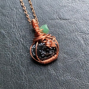 May include: A handcrafted copper wire-wrapped necklace featuring a black and white speckled stone pendant. A small green gemstone is also incorporated into the design. The necklace hangs on a delicate copper chain, creating a unique and artistic accessory.