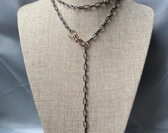 May include: A layered necklace with a dark gunmetal chain and a gold-colored clasp. The necklace is displayed on a mannequin with a neutral-colored fabric background. The chain has a rectangular link design.