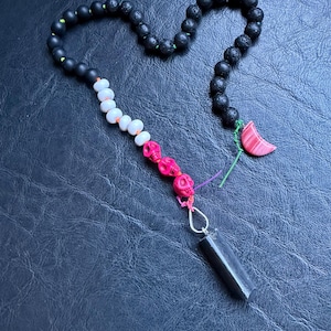 May include: A beaded necklace featuring black, white, and pink skull beads, with a black cylindrical pendant. The necklace includes black lava rock beads and a pink crescent moon charm. The beads are strung on a black cord.