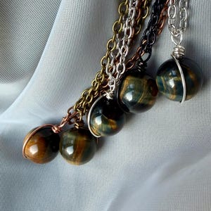 May include: A collection of gemstone pendant necklaces. Each necklace features a spherical, polished stone with a unique pattern of brown, gold, and blue hues. The stones are wrapped in wire and suspended from chains in various metallic colors.