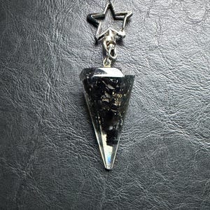 May include: A silver-toned star-shaped charm attached to a black and clear crystal pendulum. The pendulum is cone-shaped with a silver cap. The crystal has black and white inclusions. The background is a textured black surface.