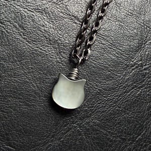 May include: A silver-toned cat pendant necklace with a dark chain. The cat pendant has a rounded shape with small, pointed ears. The chain is made of small, interlocking links.