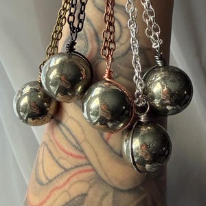 May include: Close-up of five pyrite sphere pendants on chains. The chains are in various colors: gold, black, copper, and silver. Each sphere is wrapped with wire and hangs from the chain. The background is blurred.