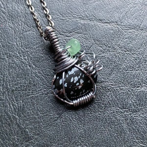 May include: A handcrafted pendant necklace featuring a black and white speckled stone wrapped in silver-colored wire. A small, light green bead is nestled within the wirework. The pendant hangs from a delicate silver chain, creating a unique and artistic accessory.