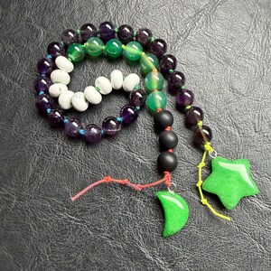 May include: A beaded necklace featuring a variety of colorful beads in shades of purple, green, white, and black. The necklace includes a green star and crescent moon pendant, and is displayed on a dark surface.