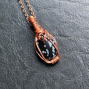 May include: A copper wire-wrapped pendant necklace featuring a black snowflake obsidian stone. The stone is oval-shaped with white speckles. The pendant hangs from a delicate copper chain, creating a unique jewelry piece.