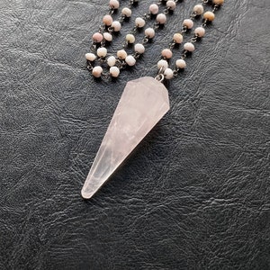 May include: A necklace featuring a faceted, light pink quartz crystal pendant. The pendant hangs from a chain of small, round, light pink and white beads. The necklace is displayed on a dark, textured surface.