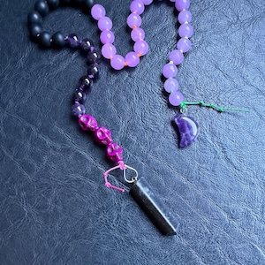 May include: A beaded necklace featuring black, purple, and lavender beads. The necklace includes skull-shaped beads in a vibrant pink hue, a dark rectangular pendant, and a crescent moon charm.