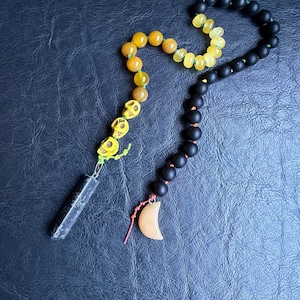 May include: A beaded necklace featuring a mix of black, yellow, and orange beads. The necklace has a pendant with a small wooden crescent moon and a black rectangular stone. The beads include skull-shaped and round beads.
